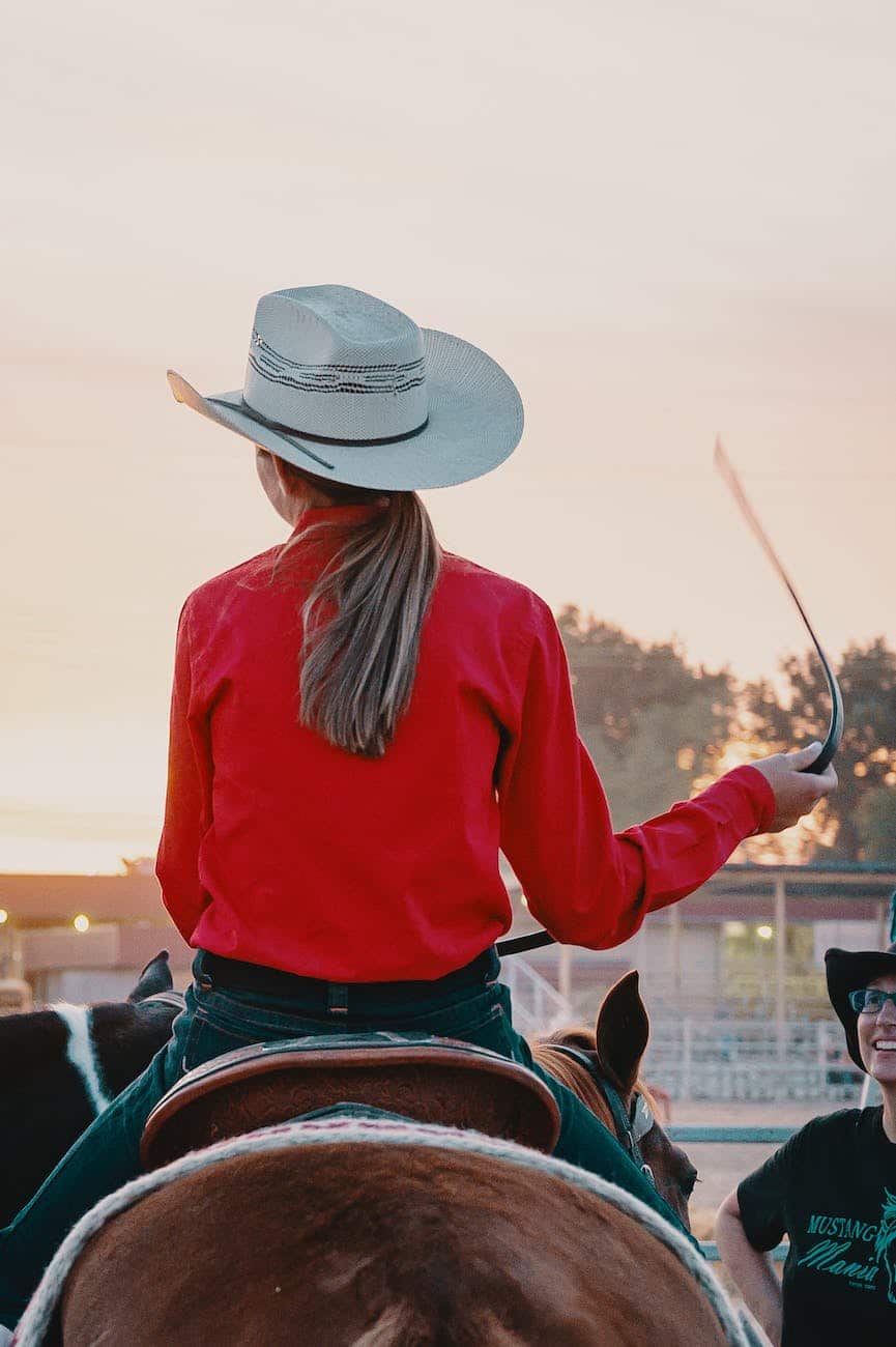 woman wearing white cowboy hat and red top riding on horse